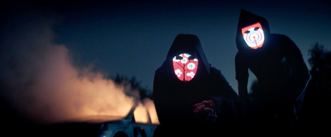 Two hooded figures with glowing LED masks at night, car burning behind