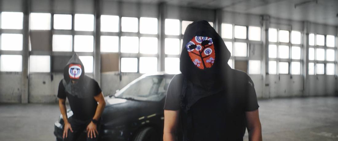 Two hooded figures with LED masks in a bright warehouse