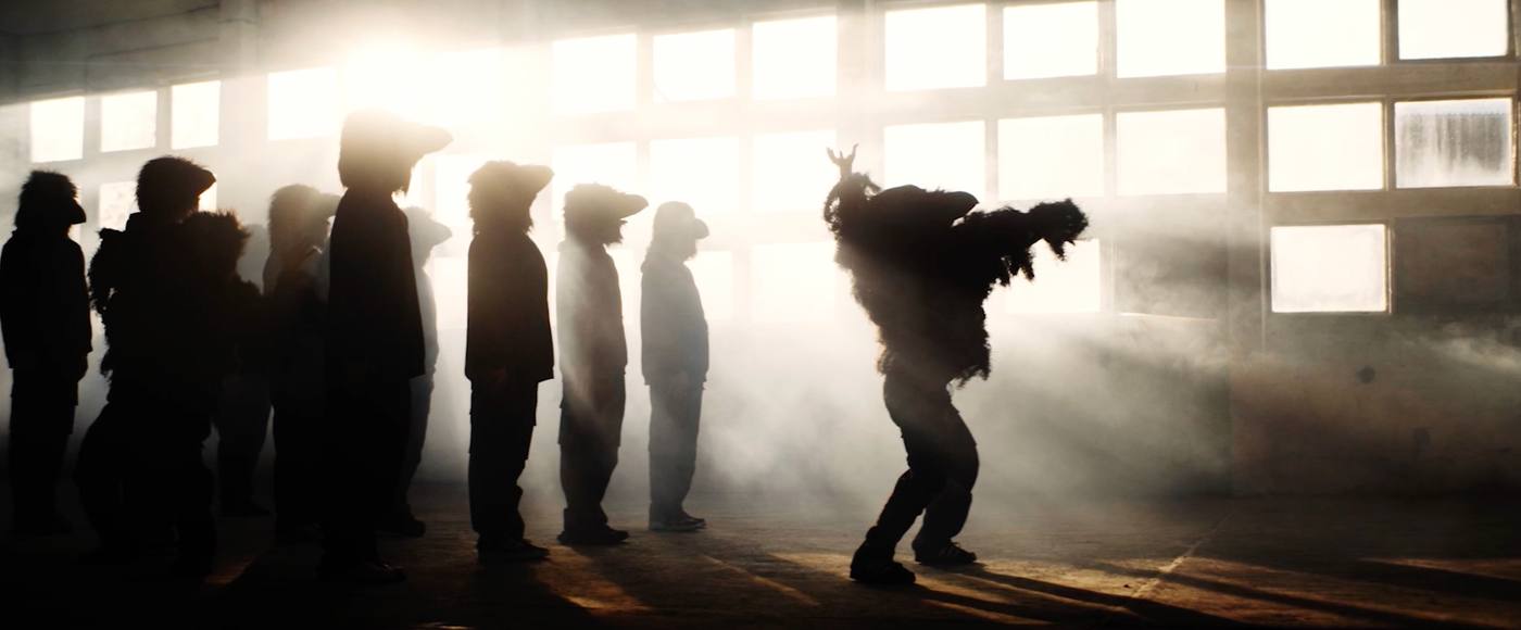 Silhouettes in fur costumes lined up in a foggy warehouse, one dancing