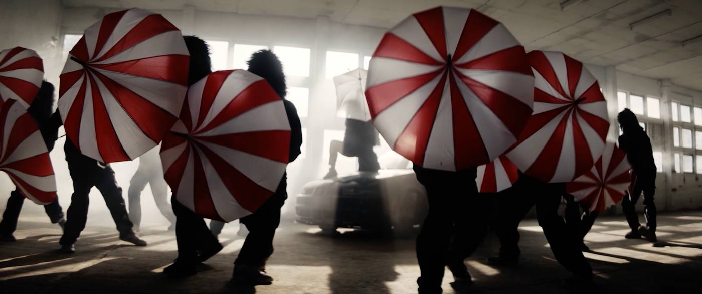 Music video still, figures in fur costumes with red-and-white umbrellas circling a car in a warehouse